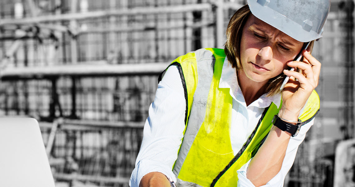 A woman wearing personal protective equpiment uses a cell phone and laptop at a construction site