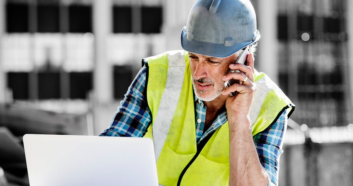 A man wearing personal protective equipment uses a cell phone and laptop on a construction site